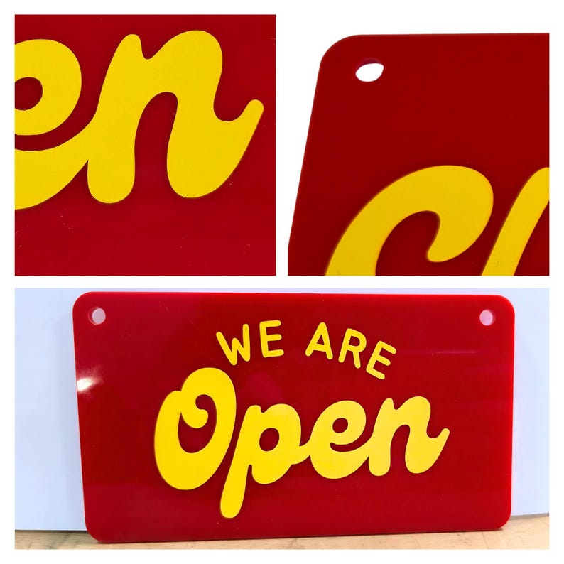 May include: A rectangular red sign with the words "WE ARE Open" in yellow, stylized lettering. The sign has rounded corners and two holes for hanging. The background is white.