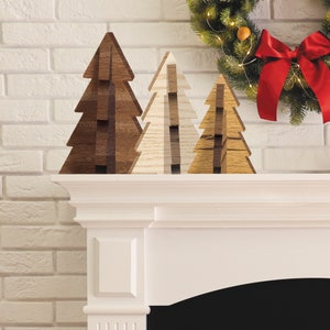 May include: Three wooden Christmas tree ornaments in different shades of brown and white, arranged on a white mantelpiece.
