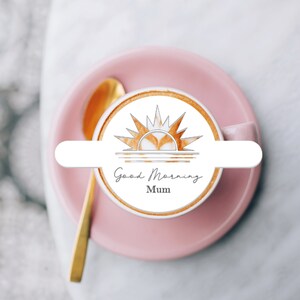 May include: A white ceramic coffee mug with a pink saucer. The coffee has a latte art design of a sun with the words "Good Morning Mum" written below it.