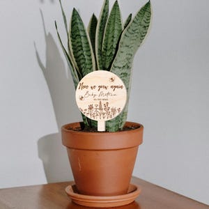 K&ouml;nnte beinhalten: Ein h&ouml;lzerner Pflanzenmarker mit dem Text "Never we grow again Baby Matthew 01/03/2023" und einem Bienen- und Blumendesign.
