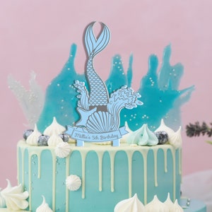 May include: A blue mermaid cake topper with a shell and seaweed, with a banner that reads "Millie's 5th Birthday".