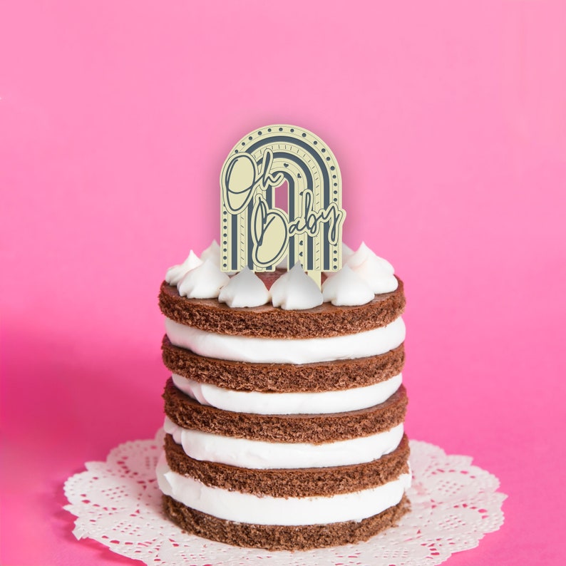 May include: A chocolate cake with white frosting is topped with a yellow and blue 'Oh Baby' cake topper shaped like a rainbow.