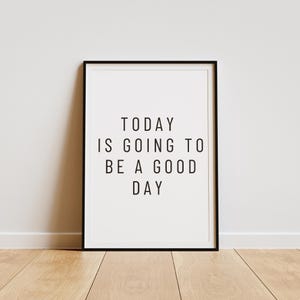 K&ouml;nnte beinhalten: Gerahmter Druck mit dem Text "TODAY IS GOING TO BE A GOOD DAY" in Schwarz, zentriert auf wei&szlig;em Hintergrund. Der Rahmen ist schwarz, und der Druck wird vor einer wei&szlig;en Wand und einem Holzboden gezeigt.