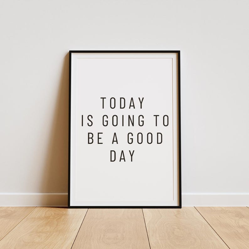 K&ouml;nnte beinhalten: Gerahmter Druck mit dem Text "TODAY IS GOING TO BE A GOOD DAY" in Schwarz, zentriert auf wei&szlig;em Hintergrund. Der Rahmen ist schwarz, und der Druck wird vor einer wei&szlig;en Wand und einem Holzboden gezeigt.