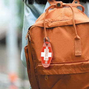 K&ouml;nnte beinhalten: Ein brauner Rucksack mit einem roten und wei&szlig;en Schl&uuml;sselanh&auml;nger mit medizinischen Informationen. Der Schl&uuml;sselanh&auml;nger ist in Form eines Motel Schl&uuml;ssels und hat ein wei&szlig;es Kreuz und den Text "MEDICAL INFO!" darauf.