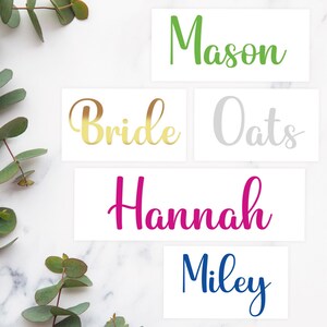 May include: Rectangular white cards with names in various colors: green "Mason", gold "Bride", gray "Oats", pink "Hannah", and blue "Miley". Eucalyptus leaves are in the background.