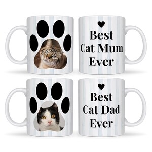 May include: Four white ceramic mugs with cat-themed designs. Two mugs feature a cat paw print with a cat's face inside. The other two mugs have the text "Best Cat Mum Ever" and "Best Cat Dad Ever" with a heart symbol. The mugs have a blue striped background.
