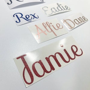 May include: A collection of glittery name decals in various colors, including red, silver, rose gold, and blue. The names are written in a cursive font and are displayed on individual white rectangular stickers. Names include Jamie, Eadie, Rex, Alfie, and Dane.