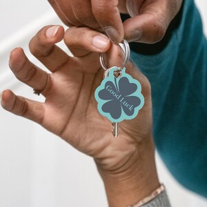 May include: A teal blue four-leaf clover keyring with a silver ring and a key attached. The clover has a dark grey outline and the text "Good Luck" written in white.
