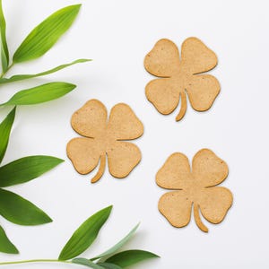 May include: Three wooden four-leaf clovers with a light brown color are arranged on a white surface, with green leaves in the background. The clovers are a symbol of good luck and are often used for St. Patrick's Day decorations.