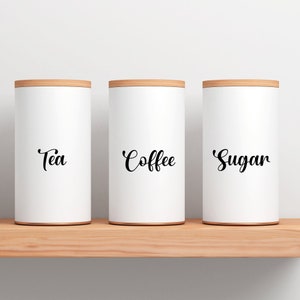 Peut inclure: Trois bo&icirc;tes blanches avec des couvercles en bois sur une &eacute;tag&egrave;re en bois. Les bo&icirc;tes sont &eacute;tiquet&eacute;es "Th&eacute;", "Caf&eacute;" et "Sucre" en &eacute;criture cursive noire.
