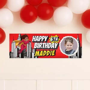 Puede incluir: Bandera roja con el texto "HAPPY 6TH BIRTHDAY MADDIE" en blanco y amarillo. Una superhero&iacute;na de dibujos animados vuela sobre un paisaje urbano. Una foto de una ni&ntilde;a est&aacute; en el lado derecho de la bandera. Globos rojos y blancos en el fondo.