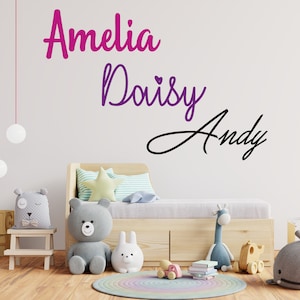 Op de afbeelding: Een witte muur met drie namen in cursief geschreven: Amelia, Daisy en Andy. De naam Amelia is geschreven in roze en paars, Daisy is geschreven in paars en Andy is geschreven in zwart. Er zijn knuffels en speelgoed voor een houten bed.