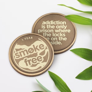 May include: Two gold-colored metal tokens. One token features the text "1 YEAR smoke free" in a white, stylized font. The other token features the text "addiction is the only prison where the locks are on the side." in a white, sans-serif font.