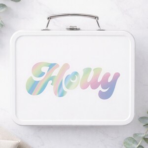 Puede incluir: Fiambrera met&aacute;lica blanca con asa plateada. La fiambrera presenta el nombre "Houy" en una fuente colorida de estilo retro con tonos pastel arco&iacute;ris. La fiambrera es rectangular.