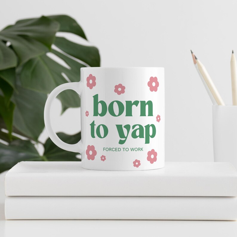 May include: White ceramic mug with green text that reads "born to yap" and the smaller text "forced to work". The mug is decorated with pink flower illustrations.