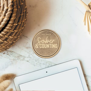 May include: A wooden round coaster with the text "100 Days Sober & Counting" in gold lettering. The coaster has a wood grain texture and a brown color.