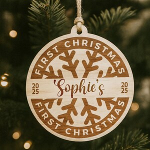 May include: A round wooden Christmas ornament with the text "FIRST CHRISTMAS" and "Sophie's" in a decorative font. The ornament has a snowflake design and the year "2025". It hangs from a natural twine.