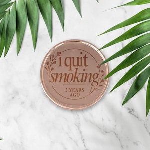 May include: A rose gold round coin with the text "I quit smoking 2 years ago" engraved on it. The coin is surrounded by a floral design.