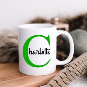 K&ouml;nnte beinhalten: Eine wei&szlig;e Keramiktasse mit einem gr&uuml;nen Buchstaben C und dem Namen Charlotte, der in Schwarz in den Buchstaben geschrieben ist. Die Tasse steht auf einer Holzoberfl&auml;che.