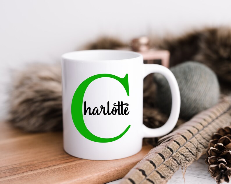 K&ouml;nnte beinhalten: Eine wei&szlig;e Keramiktasse mit einem gr&uuml;nen Buchstaben C und dem Namen Charlotte, der in Schwarz in den Buchstaben geschrieben ist. Die Tasse steht auf einer Holzoberfl&auml;che.