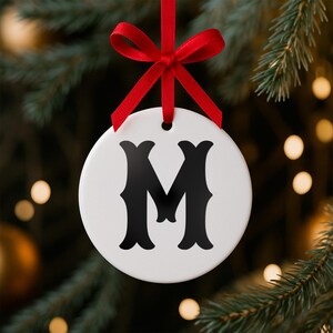 May include: A white, round Christmas ornament with a black letter "M" in a decorative font. The ornament hangs from a red ribbon tied in a bow. The background is a blurred image of a Christmas tree with bokeh lights.