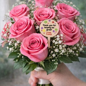 May include: A bouquet of pink roses and white baby's breath, held by a hand. A wooden sign with the text "Thank you for helping me grow! From Penny" is attached to the bouquet. The flowers are tied with a white ribbon.