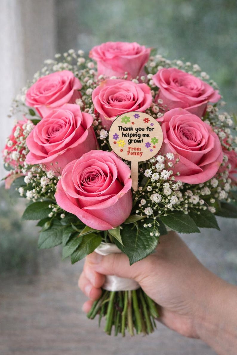 May include: A bouquet of pink roses and white baby's breath, held by a hand. A wooden sign with the text "Thank you for helping me grow! From Penny" is attached to the bouquet. The flowers are tied with a white ribbon.