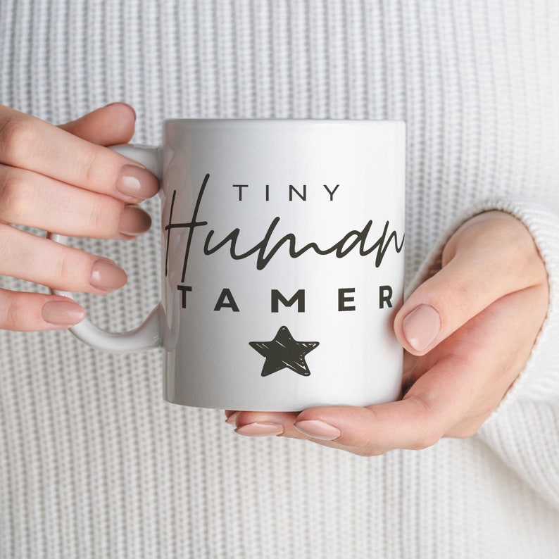 May include: White ceramic mug with black text that reads "Tiny Human Tamer" and a black star. The mug is being held by a person.