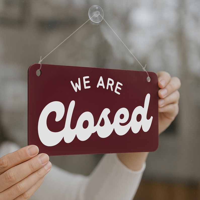 May include: A rectangular maroon sign with the words "WE ARE Closed" in white, hanging from a clear suction cup. The sign is held up by a thin string, indicating a business is currently not open.