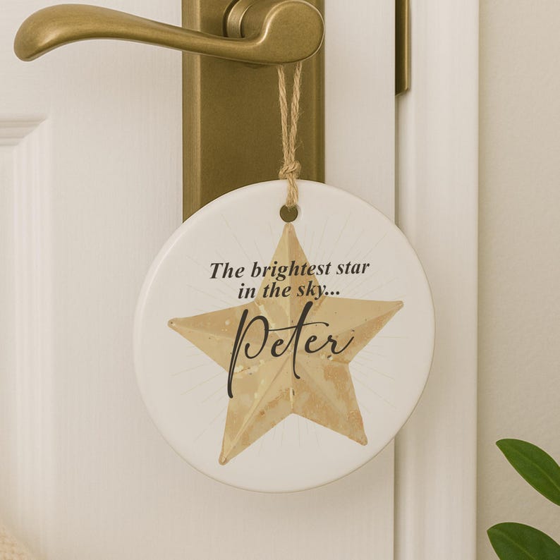 May include: A round, white ceramic ornament with a gold star design and the name "Peter" in script. The text "The brightest star in the sky..." is above the star. A twine loop is attached for hanging.