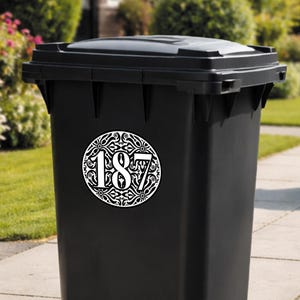 May include: A black trash bin with a decorative white and black address number sticker. The sticker features the number 187 in a stylized font, surrounded by an ornate circular design. The bin is outdoors, with greenery in the background.