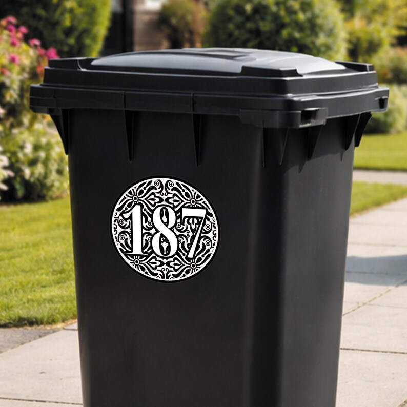 May include: A black trash bin with a decorative white and black address number sticker. The sticker features the number 187 in a stylized font, surrounded by an ornate circular design. The bin is outdoors, with greenery in the background.