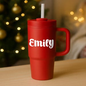 May include: A red tumbler with a handle and a white straw. The word "Emily" is printed in white script on the front of the tumbler. The tumbler is sitting on a wooden surface with a blurred Christmas tree in the background.