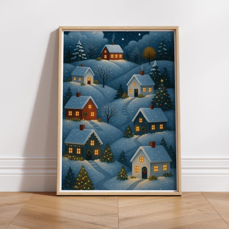 May include: A framed print of a winter village scene. Snow-covered hills feature several houses with lit windows and chimneys. The artwork has a soft, painterly style, with a night sky filled with stars and falling snow. Christmas trees are decorated with lights.