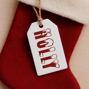 以下が含まれることがあります： 赤い文字で「HOLLY」と書かれた白いギフトタグが、茶色の紐から吊り下げられています。タグは、白いカフが付いた赤いクリスマスストッキングの上に配置されています。文字は、お祭り的で滴るようなデザインです。