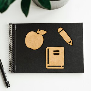 K&ouml;nnte beinhalten: Ein schwarzes Notizbuch mit Spiralbindung und drei Holzausschnitten: ein Apfel, ein Buch und ein Bleistift. Die Ausschnitte sind hellbraun.