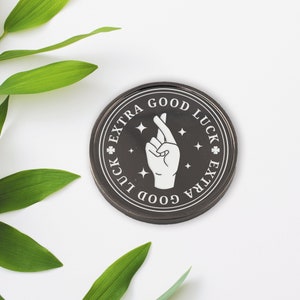 May include: A black round coin with a white design of a hand with crossed fingers. The coin has the text "EXTRA GOOD LUCK EXTRA" around the edge.