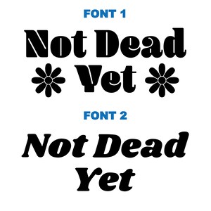 Op de afbeelding: Twee versies van de zin "Not Dead Yet" in zwarte tekst op een witte achtergrond. De eerste versie is in een vetgedrukt, blokkerig lettertype met gestileerde madeliefjes aan weerszijden van het woord "Yet". De tweede versie is in een meer casual, afgerond lettertype.