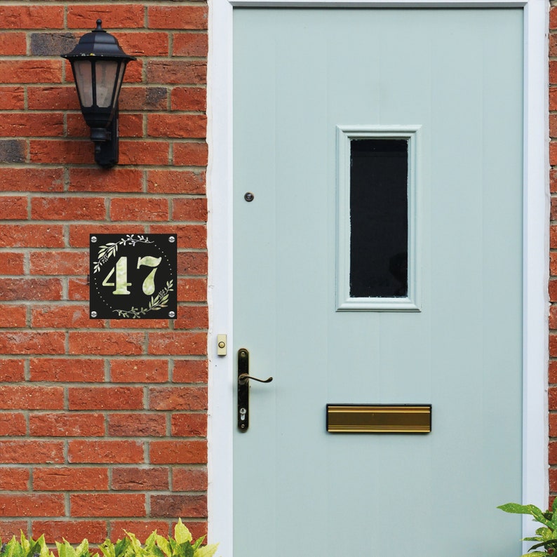 May include: A black house number sign with a white background and the number 47 in the center. The sign is surrounded by a green floral design. The sign is mounted on a brick wall next to a light blue door with a gold mailbox.