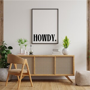 Op de afbeelding: Een zwart ingelijste poster met het woord "HOWDY." gedrukt in wit op een witte achtergrond. De poster hangt aan een witte muur boven een houten kast met een geweven rotanfront. De kast staat op een houten vloer. Er staan twee planten in potten op de kast.