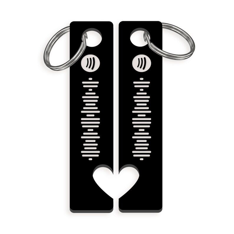 May include: Black metal keychain with a heart-shaped cutout. The keychain features a white sound wave design and a silver ring.