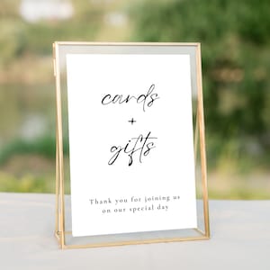May include: A gold-coloured framed sign displaying the words "cards + gifts" in cursive script. The text "Thank you for joining us on our special day" is below. The sign is on a white surface with a blurred green backdrop.