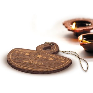 K&ouml;nnte beinhalten: Ein Holz-Diya-Anh&auml;nger mit einer Schnur zum Aufh&auml;ngen. Der Diya ist mit Sternen verziert und das Wort "Diya" ist auf der Vorderseite eingraviert.