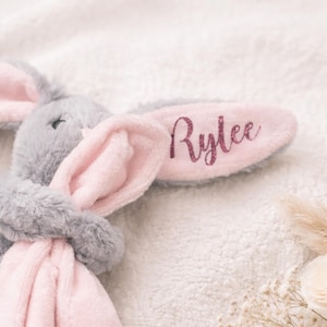 May include: A soft grey bunny toy with pink inner ears and a pink belly. The right ear features the name "Rylee" in glitter lettering. The bunny is resting on a fluffy, white, textured surface.
