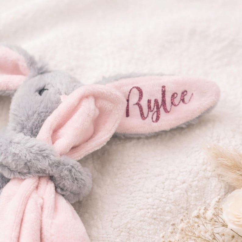May include: A soft grey bunny toy with pink inner ears and a pink belly. The right ear features the name "Rylee" in glitter lettering. The bunny is resting on a fluffy, white, textured surface.