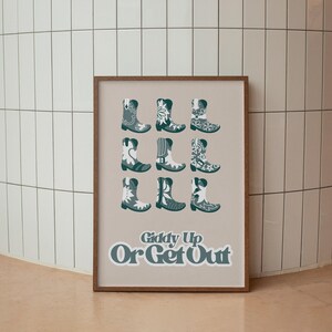 May include: A beige poster with a white background featuring nine pairs of teal cowboy boots with white floral patterns. The poster has the text "Giddy Up Or Get Out" in white letters with a teal outline.
