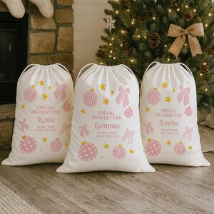 Puede incluir: Tres bolsas de regalo navide&ntilde;as blancas con cierres de cord&oacute;n. Cada bolsa presenta dise&ntilde;os festivos rosas y amarillos, incluyendo adornos, lazos y estrellas. El texto en las bolsas dice "Special Delivery For:" seguido de un nombre y "Do Not Open Until the 25th."