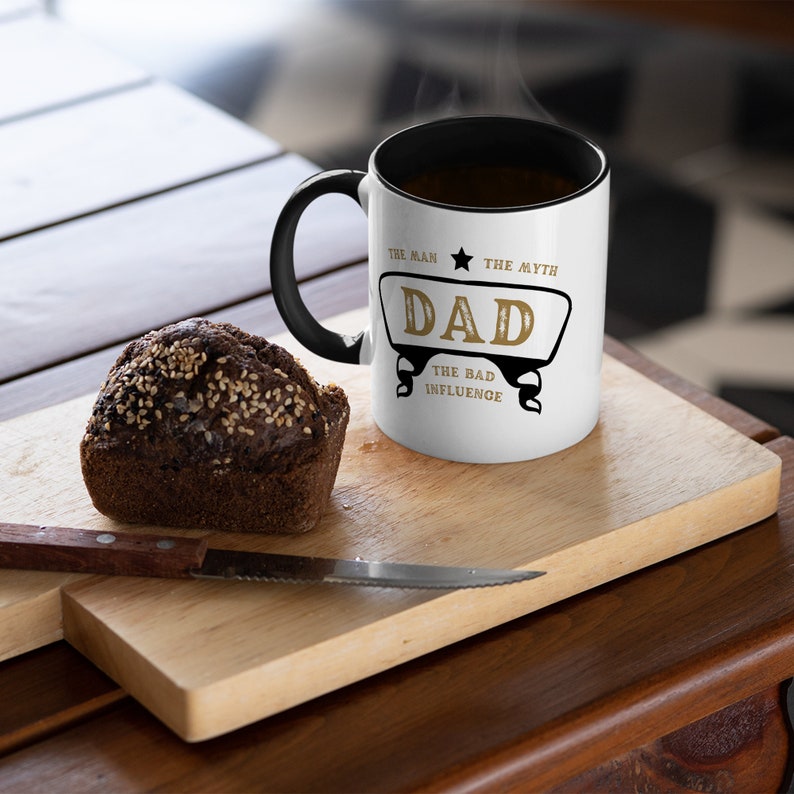 May include: White coffee mug with a black handle. The mug has black text that reads "The Man, The Myth, Dad, The Bad Influence" with a gold star and a gold banner.