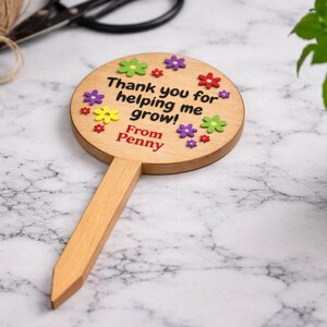 May include: Wooden garden stake with a round top and a pointed base. The top is decorated with colorful flower accents and the text "Thank you for helping me grow! From Penny". A plant in a pot is in the background.